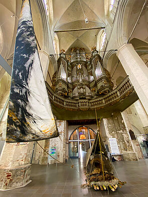 Im Kirchenschiff steht ein Boot. Gebaut aus Asten. Eine Plane spannt sich wie ein Zeltdach darüber. Im Inneren liegen Felle und Decken. Daneben hängt ein überdimensionales Segel.