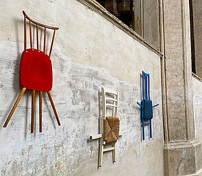 An der Wand hängen drei Stühle. Sitzfläche, Lehne und Stuhlbeine wurden neu zusammengesetzt. Jetzt sind die Stühle ganz flach.