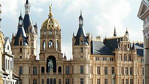 Außenaufnahme vom Schweriner Schloss. Der Ausschnitt startet im dritten Stock, erfasst das Reiterstandbild und mehrere Türme und Türmchen. Links und rechts deuten sich Fassaden historischer Gebäude aus der Schlossstraße an - so, als grenzten sie unmittelbar ans Schloss.