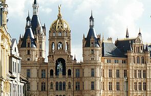 Außenaufnahme vom Schweriner Schloss. Der Ausschnitt startet im dritten Stock, erfasst das Reiterstandbild und mehrere Türme und Türmchen. Links und rechts deuten sich Fassaden historischer Gebäude aus der Schlossstraße an - so, als grenzten sie unmittelbar ans Schloss.