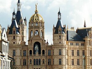 Außenaufnahme vom Schweriner Schloss. Der Ausschnitt startet im dritten Stock, erfasst das Reiterstandbild und mehrere Türme und Türmchen. Links und rechts deuten sich Fassaden historischer Gebäude aus der Schlossstraße an - so, als grenzten sie unmittelbar ans Schloss.