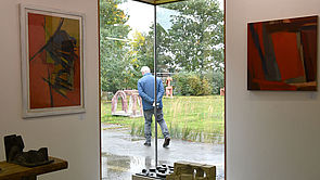Eine Hausecke aus Glas. Im Inneren hängen Bilder an der Wand. Außen läuft der Künstler entlang. 