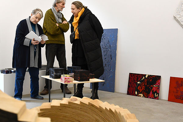Broder Burow zwischen zwei Frauen in einer Galerie. Im Hintergrund lehnen Malereien an einer Wand