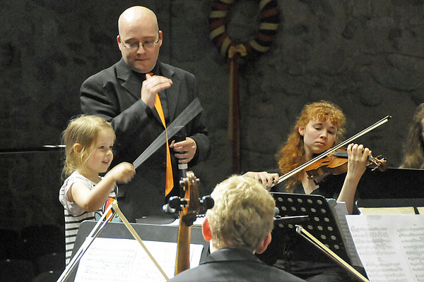 Ein Kind hält einen Taktstock in der Hand und dirigiert. Neben ihr steht der Dirigent. Eine junge Frau spielt Geige.