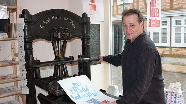 Hans-Hilmar Koch steht vor einer schwarzen Druckmaschine und hält ein bedrucktes Blatt in der Hand. 