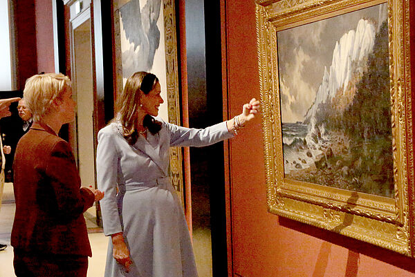 Zwei Frauen betrachten ein Ölgemälde. Die Malerei zeigt eine Gebirgslandschaft.