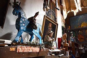 Im Atelier steht ein großes blaues Holzpferd. Daneben hängen Marionetten. Marionetten sind Puppen an Fäden. Dahinter hängen viele bunte Gemälde.