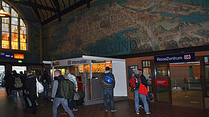 Im Bahnhof Stralsund befindet sich ein großes Gemälde auf der Wand. In der Halle befinden sich viele Menschen.