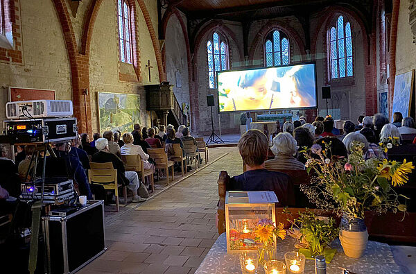 Eine Kirche mit voll besetzten Sitzreihen. Vor dem Altar steht eine große Leinwand, auf der ein Film läuft.