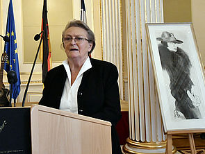 Inge Jastram steht an einem Rednerpult und spricht. Hinter ihr steht eine Zeichnung. Darauf: eine Frau mit Hut und langem Kleid.