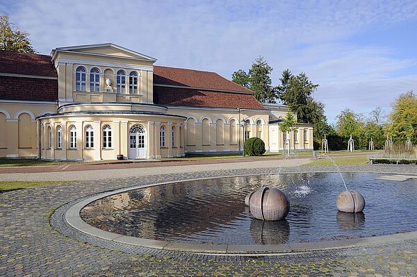 Ein historisches Gebäude im Schlossgarten. Davor liegen zwei Stein in einem Becken mit flachem Wasser. Aus den Steinen kommen Wasserstrahlen.