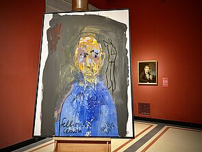 Ein Porträt steht auf einer Staffelei vor einer roten Wand.