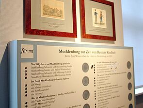 An der Wand hängt ein Wissensquiz. Darüber hängen Bilderrahmen mit Zeichnungen.