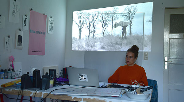 Stefanie Rübensaal sitzt an einem Schreibtisch. Darauf befinden sich Zettel, Boxen, Kabel und ein Laptop. An der Wand - ein Videobild von einem Mann zwischen Dünen. 