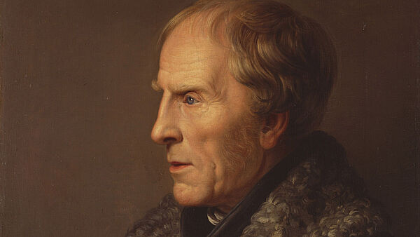 Caspar David Friedrich im Seiteprofil vor einem braunen Hintergrund. Er ist um die 60 Jahre alt, trägt helles Haar und einen Umhang mit Fellbesatz.