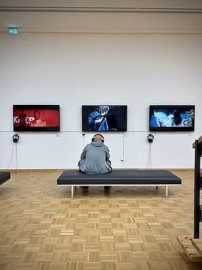 Ein Mann sitzt vor einer Wand mit drei Bildschirmen und schaut Videos.