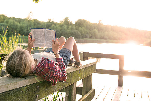 Ein Mädchen liegt an einem See. Sie liegt an einem Steg, auf einem Holzbalken und liest ein Buch.