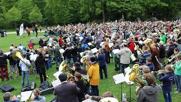 Auf einer Wiese stehen, halbrund aufgestellt, mehrere hundert Posaunisten und spielen ihr Instrument.