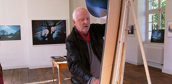 Volkmar Herre steht an einer Staffelei. Im Hintergrund hängen Bilder an der Wand.