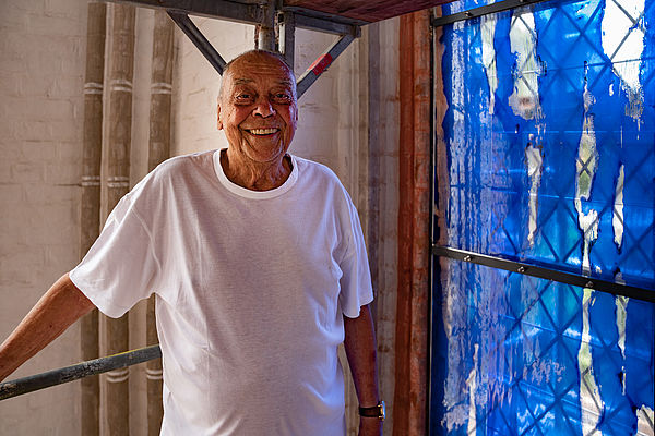 Günther Uecker steht auf einem Baugerüst vor einem blauen Kirchenfenster, das er gestaltet hat.