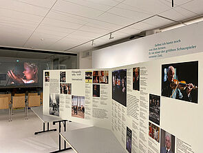 Auf zwei Tischen steht eine große Infotafel. Im Hintergrund hängt eine Leinwand. Darauf läuft ein Film mit Armin Mueller-Stahl.