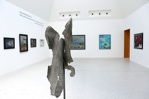Blick in einen Ausstellungsraum. Vorn steht eine Skulptur, an der Wand hängen Bilder.