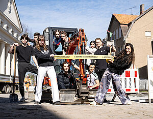 Zehn junge Leute. Sie in oder stehen an einem Bagger. Der Bagger steht auf einer Baustelle. Links und rechts stehen Häuser. Zwei Leute halten ein gelbes Absperrband. Darauf steht: Achtung Starkstromkabel!