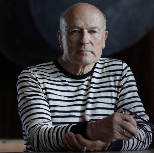 Volker Schlöndorff im Porträt. Er sitzt im geringelten Pullover vor einer dunklen Wand. 