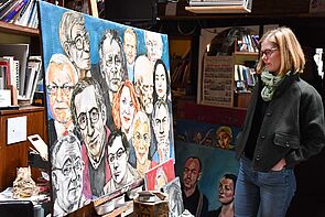 Eine Frau schaut sich ein Gemälde an. Das Gemälde zeigt viele Gesichter. Das Gemälde hat Hans W. Scheibner gemalt.