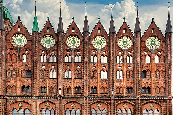 Die Schaufassade des alten Rathauses ragt mit ihren Backsteinen in den Himmel. Unten deuten sich große Glasfenster an. Über ihnen: Wappen benachbarter Hansestädte. Die Fenster darüber sind entweder mit Backsteinen verschlossen oder ohne Glas offen. In den spitz zulaufenden Giebeln befinden sich kirchenuhrgroße Ornamente. 