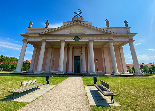 Ein tempelartiges Gebäude. Vor dem Eingang stehen acht Säulen. Auf dem Dach verteilen sich vier Statuen. Auf der Spitze des Daches stehen ein P und ein X. Ein Schotterweg führt zur Kirche. Links und rechts davon wächst Wiese und stehen Bänke. 