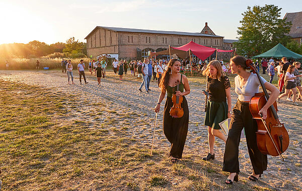 Drei junge Frauen laufen mit Instrumenten über eine Wiese. Im Hintergrund gehen Festivalbesucherinnen und Festivalbesucher über das Gelände.