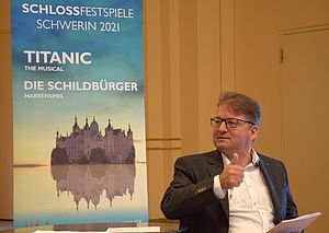 Lars Tietje sitzt neben dem Titanic-Plakat für die nächsten Schlossfestspiele und spricht.