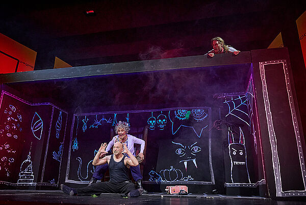 Der Teufel mit den drei goldenen Haaren sitzt auf der Bühne. Dahinter steht eine Frau. Von oben schaut ein junger Mann auf die beiden.