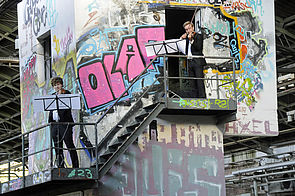 Zwei Geiger stehen auf einer Außentreppe und spielen. Hinter ihnen: ein Graffiti besprühtes Gebäude.