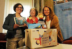 Drei Frauen stehen nebeneinander an einem Tisch. Vor ihnen steht eine Holzkiste. Die Frauen haben Bücher und Handpuppen in der Hand. Rechts steht Kulturministerin Bettina Martin.