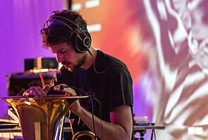 Ein junger Mann trägt Kopfhörer. Er sitzt vor einer Tuba und bewegt seine Hände auf der Öffnung des Instruments. Im Hintergrund: eine bunte Projektion.