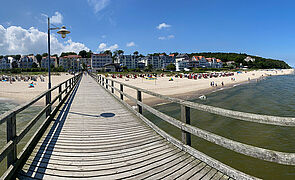 Eine Seebrücke. Links und rechts befindet sich Wasser. Am Ende des Weges kommt der Strand. Dahinter stehen Häuser.