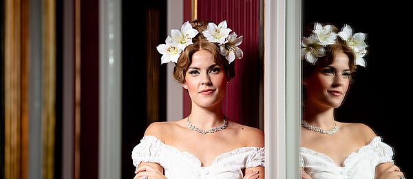 Eine Frau mit einem weißen Kleid und Blumen im Haar. Sie spiegelt sich in einem Glas.