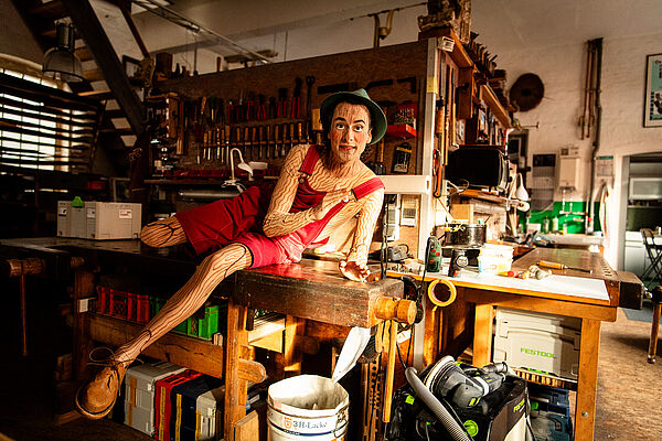 Ein Mann in einem Holzpuppenkostüm sitzt auf einer Werkbank.