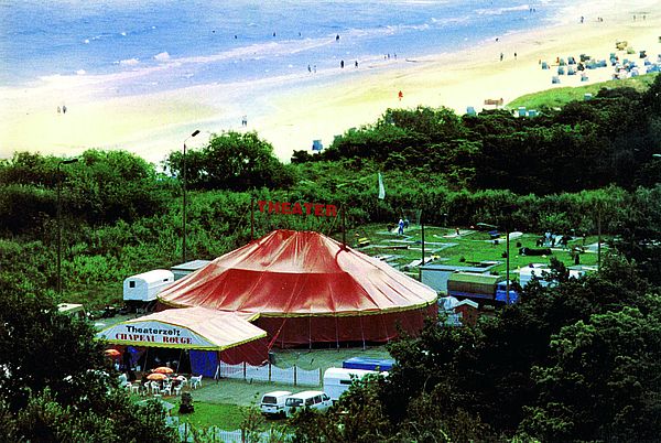 Auf einer Wiese steht ein rundes, rotes Zelt. Über seinem Dach steht das Wort "Theater". Vor dem Eingang stehen Tische und Stühle.