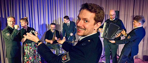 Ein Mann und eine Frau stehen vor einem Mikrofon und singen. Fünf Männer spielen Instrumente. Vor allen steht ein Dirigent. Einige der Männer tragen eine Polizeiuniform. 