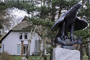 Ein weißes, restgedecktes Haus, umgeben von Wiese und Bäumen. Im Vordergrund steht auf einem weißen Sockel die Büste eines Greifvogels, der einen Fisch in seinen Krallen hält.