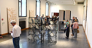 In einem Ausstellungsraum stehen Exponate. Besucherinnen und Besucher gehen durch den Raum.