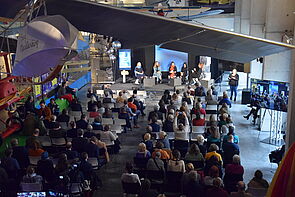 Eine Halle im Museum. Auf dem Podium wird diskutiert. Davor sitzen viele Zuschauerinnen und Zuschauer. 