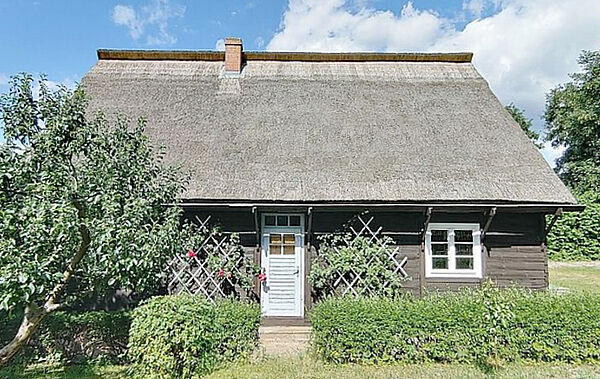 Ein Holzhaus mit Reetdach. Vor dem Haus stehen Bäume und Sträucher.