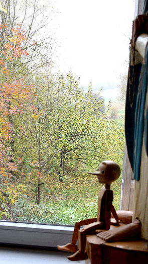 Pinocchio sitzt als Holzfigur vor dem Fenster. Draußen stehen Bäume.