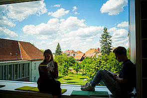 Ein großes Fenster. Auf der Fensterbank sitzen zwei Jugendliche und lesen. Dahinter stehen der Anbau des Hauses und Bäume im Garten.