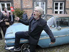 Jes Holtsø tanzt mit einem dänischen Fähnchen in der Hand vor einem blauen Oldtimer.