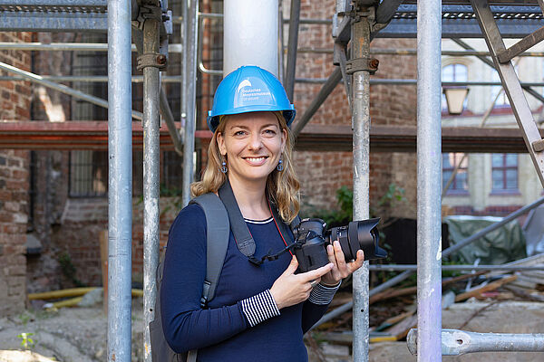 Eine blonde Frau mit einem Bauarbeiterhelm und einer Kamera in der Hand.